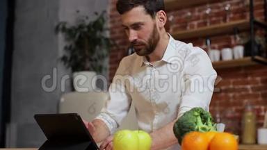 男人在家庭厨房的<strong>平板</strong>电脑上浏览。 英俊的年轻人在<strong>平板</strong>电脑上浏览，<strong>微笑</strong>着快乐。 一个男人吃